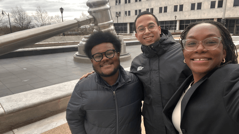Three law students participating in the Capital Habeas Corpus Externship smiling in front of a large gavel sculpture outside a courthouse.